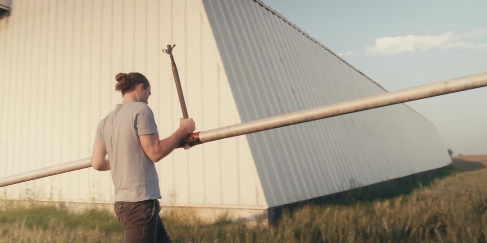 A man walks into the background carrying a long irrigation pipe next to a farm building towards a farm tractor and open fields