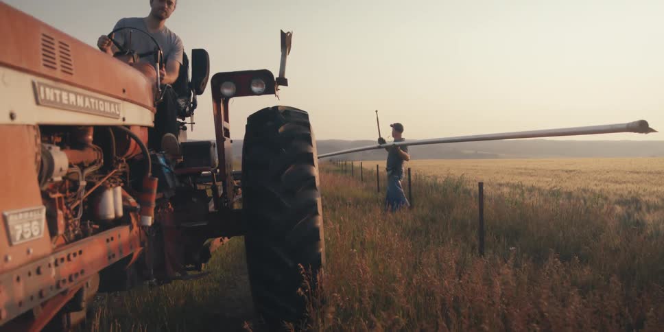 In a rural setting a man sits atop a vintage tractor while another carries a long pipe along a fence line in a sunlit field