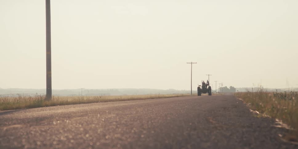 Seen from a distance down a rural road a tractor driven by a farmer travels toward camera through the hazy landscape