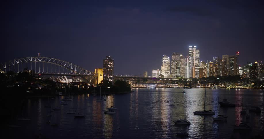 A nighttime shot shows the sydney harbour bridge illuminated against a dark sky with the city skyline and harbor lights reflecting on the water