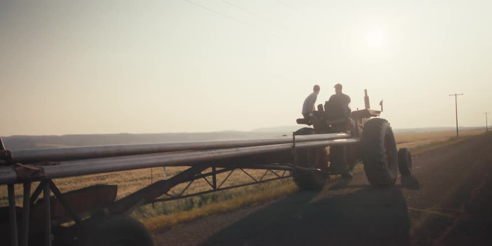 A tractor pulls a trailer loaded with pipes along a rural road under a hazy sky