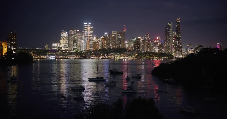 A nighttime cityscape with illuminated skyscrapers is seen across a bay where sailboats are docked the lights reflecting on the rippling surface with a tugboat in the scene