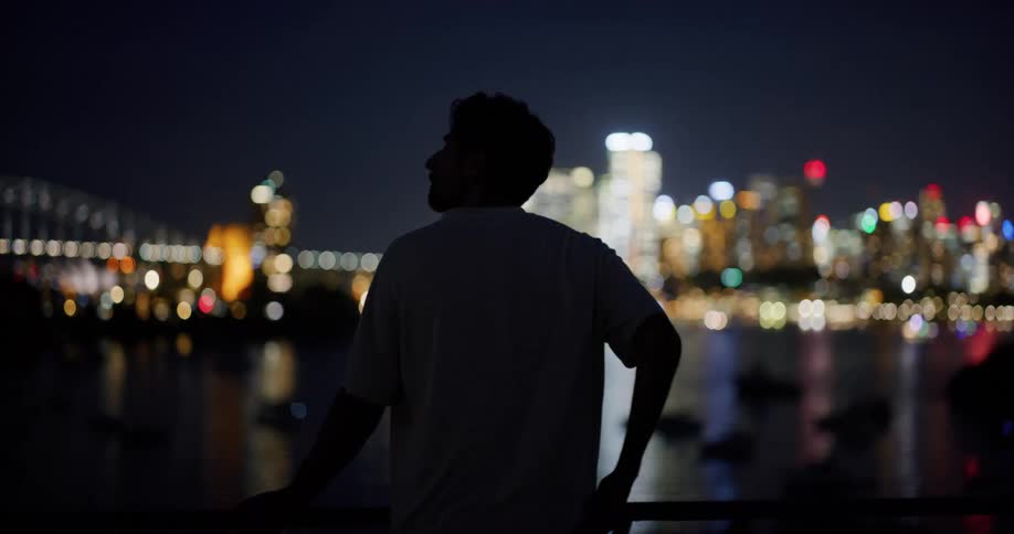 A silhouetted figure stands facing away with a city skyline illuminated by night lights in the blurred background