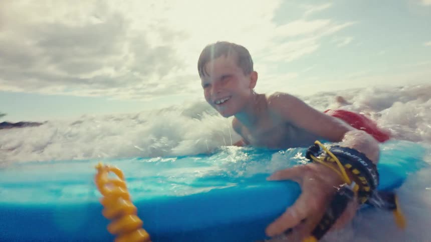 A boy rides a blue boogie board in the foamy surf of a wave with a bright sky and clouds above