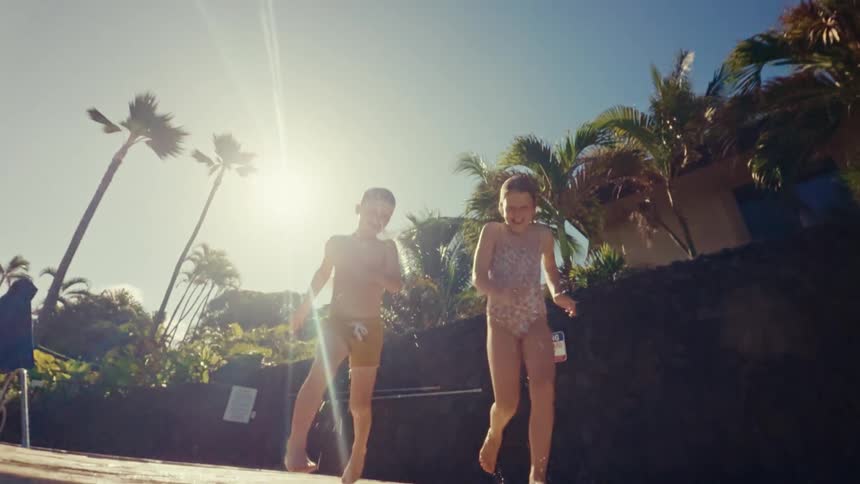 A boy and girl in swimsuits jump over the camera into a pool at a resort on a sunny day