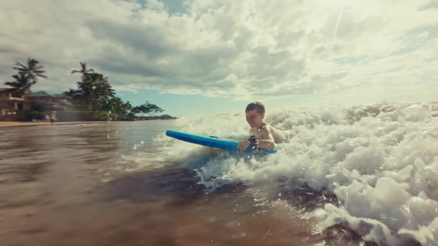 A young boy on a blue boogie board rides the foamy surf of a wave as it breaks on a sunny beach with palm trees and other people visible in the background
