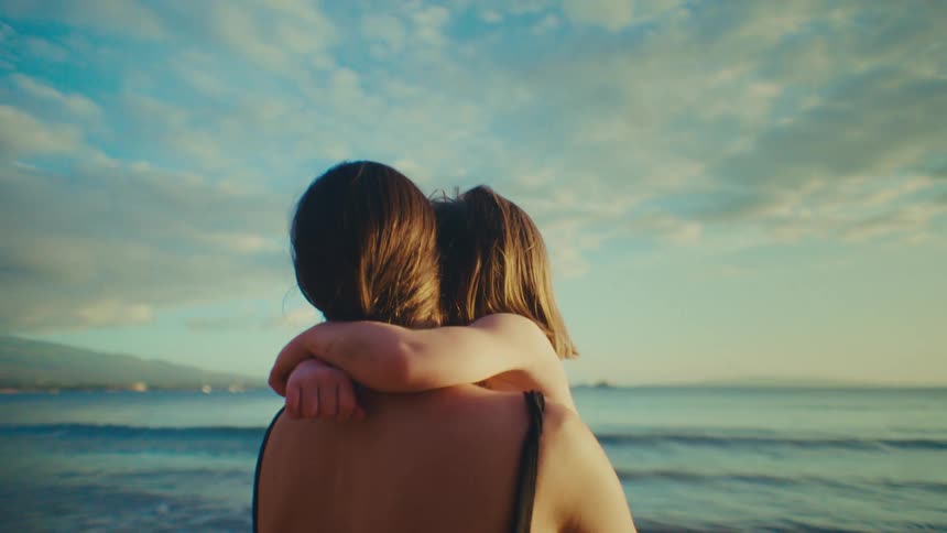 A mother holds her young daughter while looking out from a beach at dusk