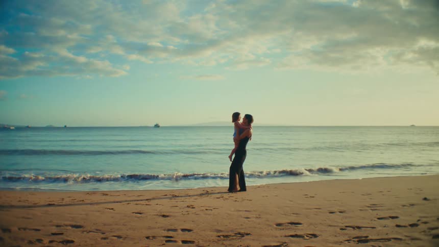 The mother is carrying the little girl up the beach in front of the waves crashing onto the sand
