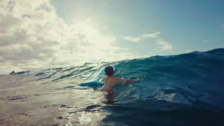 A boy riding a blue boogie board on a large ocean wave with a bright sky and clouds above
