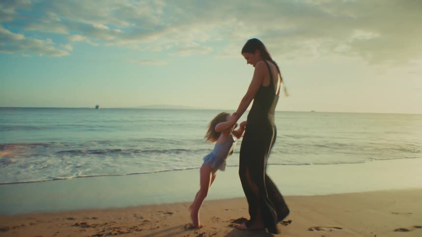 The woman in the black dress is playing and spinning with the little girl in the blue bathing suit on the beach in the sand next to the waves crashing on the beach