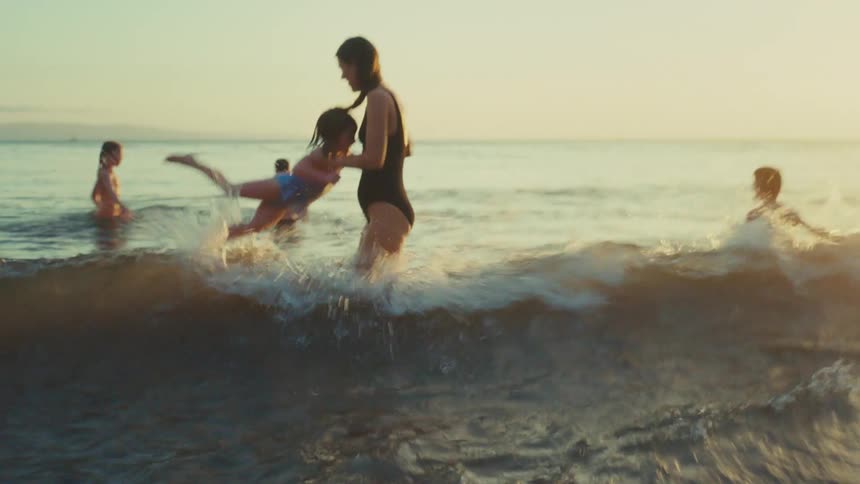 A mother joyfully plays in the ocean at sunset lifting and spinning her child in the water while another child looks on