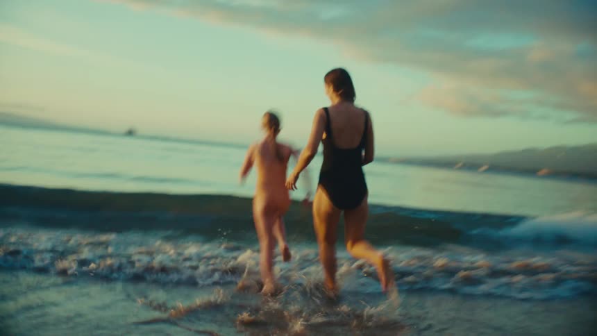 A mother and her children wearing swimwear run joyfully along the beach at sunset with the coastline in the background