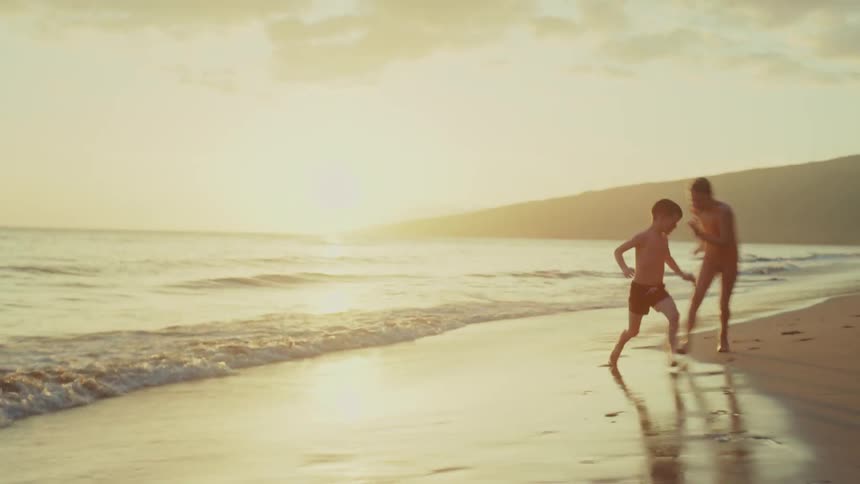 Siblings chase each other while playing on the beach near sunset on a cloudy day