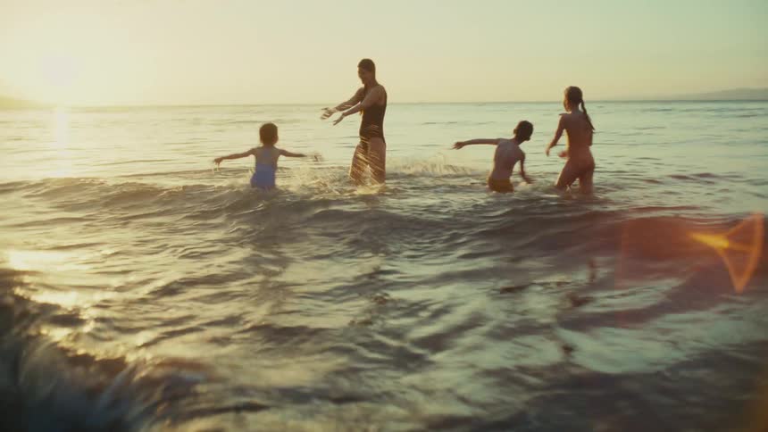 The mother is playing with her kids in the water and she picks up the little girl and spins her while the other children and laughing and splashing
