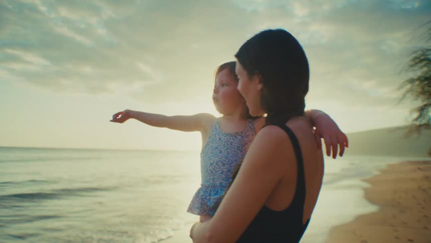 The little girl is being held on the beach beside the water by her mother and they are laughing and talking
