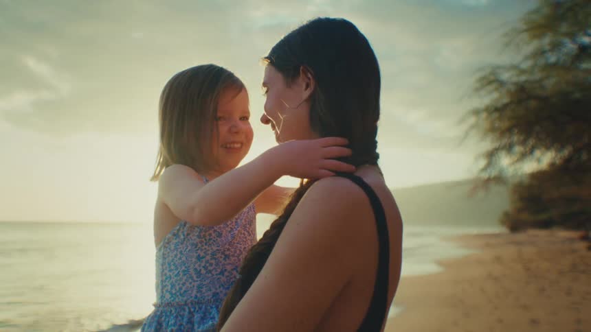 A young daughter with light hair smiles up at her mother with dark hair on a beach against a bright outdoor background