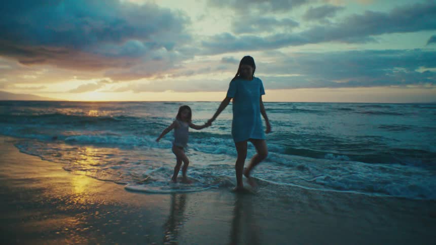 A silhouetted mother and child walk along a beach at dusk with the ocean waves lapping at their feet