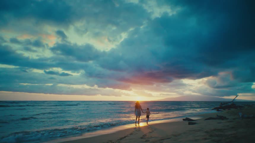 A mother holds her toddler sons hand as they walk along the beach at sunset