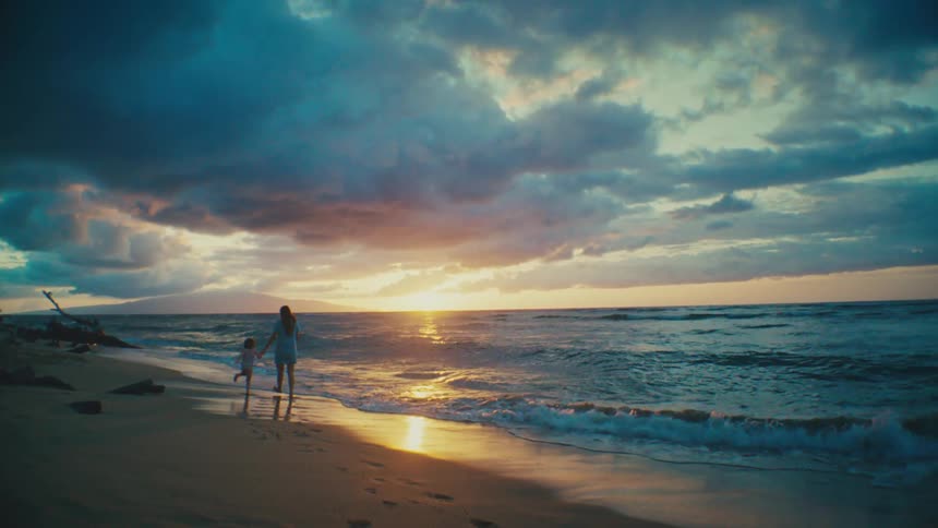 Mother and daughter holding hands while walking on the beach near the water at sunset