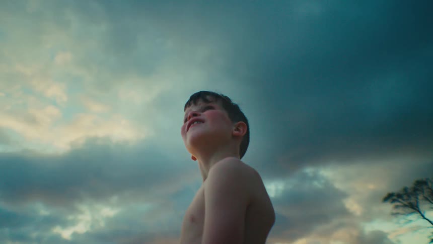 A young boy with dark hair is captured in a medium shot from a low angle revealing his bare torso and side profile as he gazes upwards towards a dramatic sky filled with dark textured clouds