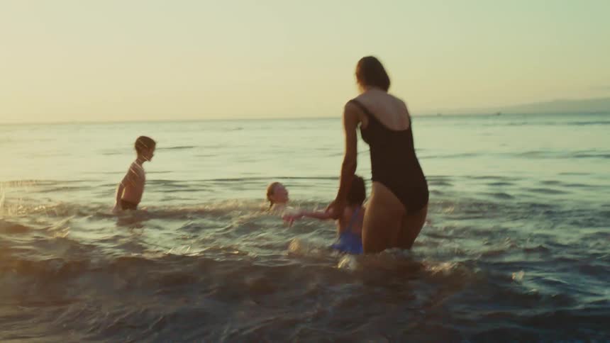 The mother is playing with her three kids in the water and she is holding the little girl under the arms and spinning her