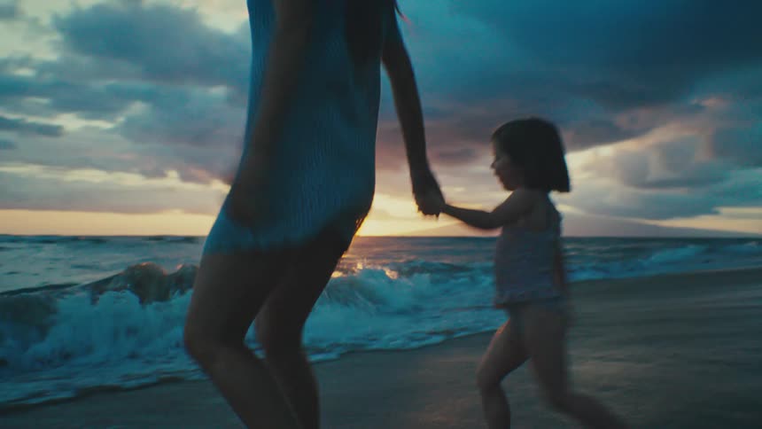 Mother and daughter playfully run towards and away from the surf while the sun sits on the horizon