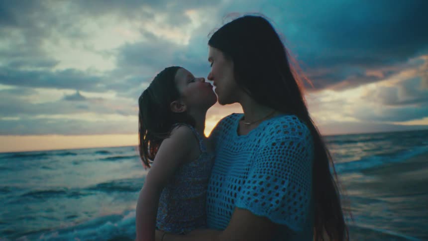 A mother and her young daughter share a kiss on the beach at sunset with the ocean and a colorful sky in the background