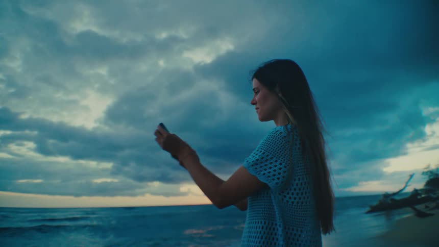 A woman with long dark hair wearing a light blue patterned shirt stands on a beach at dusk holding a phone in her hands against a backdrop of a dramatic sky with dark clouds and a sliver of sunset