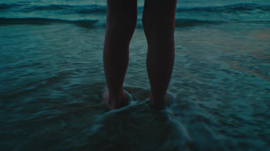 A childs bare legs and feet standing on a sandy beach with gentle waves washing ashore in the background