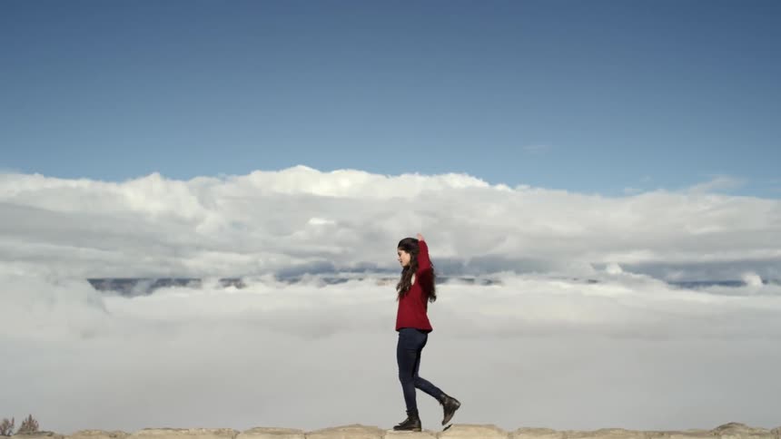 Brunette woman wearing a red jacket walking across the top of a wall with clouds in the background