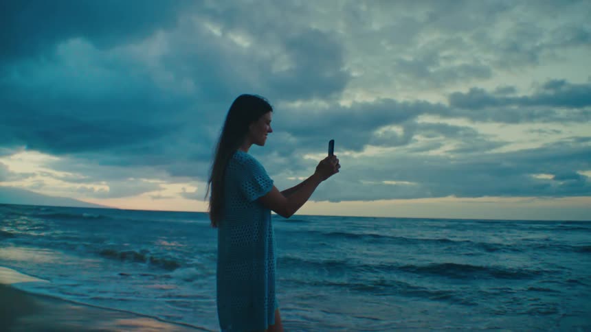 Woman standing at shoreline taking sunset pictures on cell phone at the beach