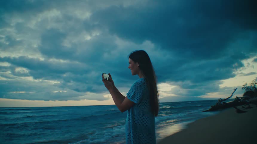 A woman with long dark hair wearing a light blue patterned top takes a photo on the beach with her phone against a dramatic backdrop of a cloudy sky and ocean