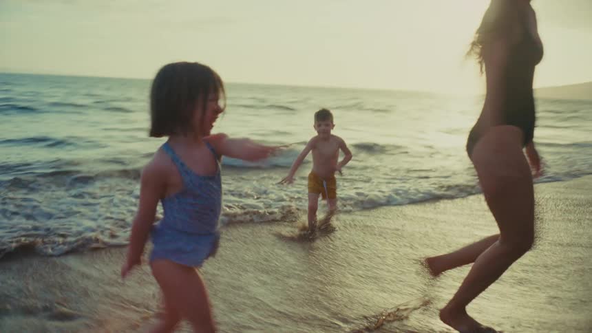 A mother and her children wearing swimwear run joyfully along the beach at sunset with the coastline in the background