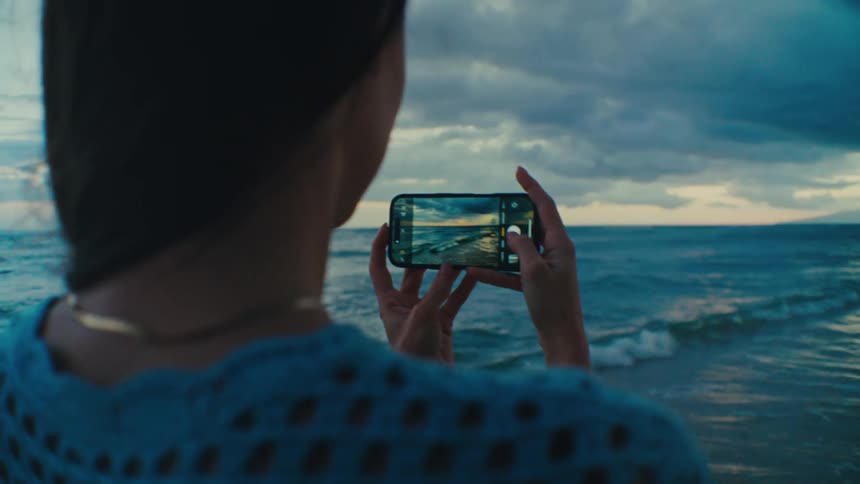 Woman taking sunset pictures on cell phone next to the shoreline at the beach
