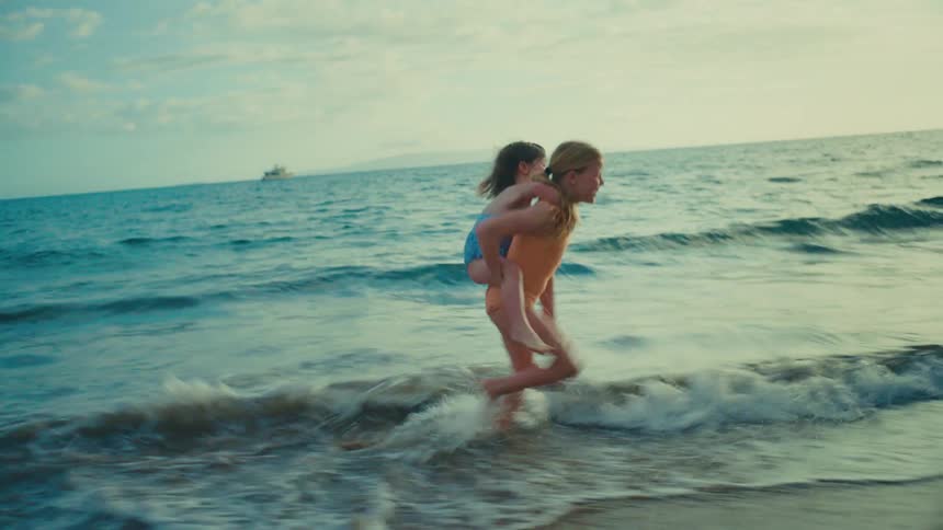 The older girl is carrying the younger girl on her back as she runs through the sand and the surf