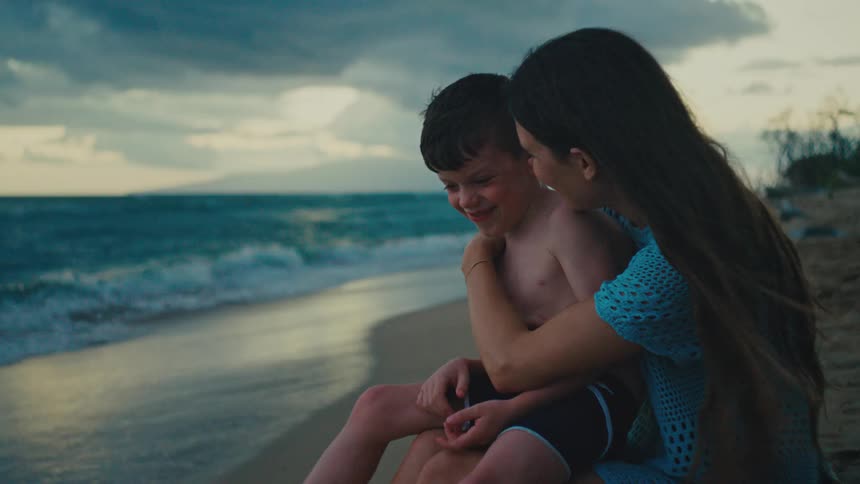 A mother lovingly embraces her young son on a beach at dusk with the ocean waves visible in the background