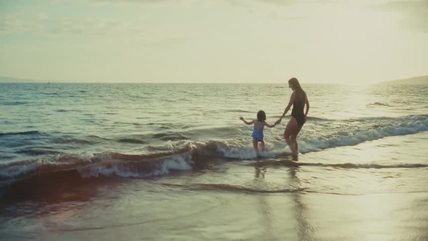 A woman and a young child walk hand in hand through shallow ocean waves on a sunny beach with land visible in the distance