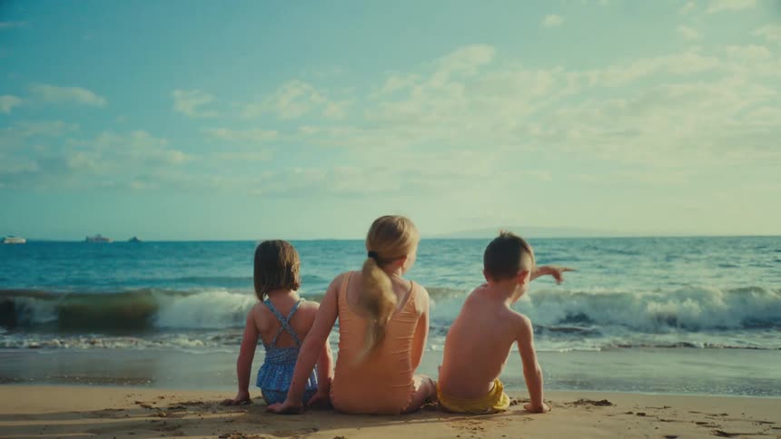Kids sitting on the beach point out things to each other while watching the ocean on a cloudy day