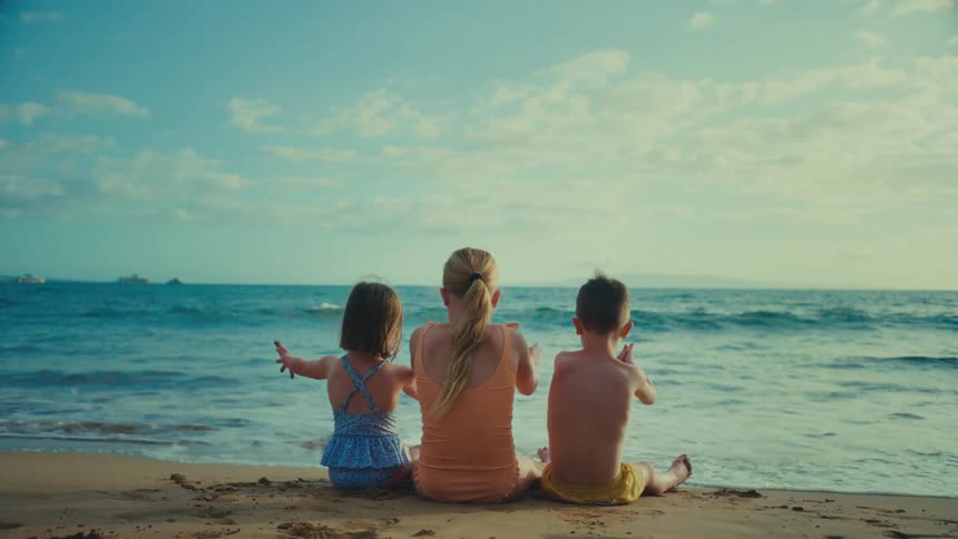 Three children two girls and a boy sit with their backs to the viewer on a sandy beach facing the ocean under a partly cloudy sky
