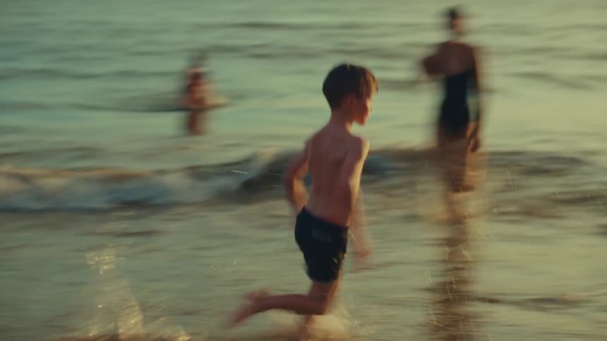 The little boy runs through the surf while his mother and brother are watching him from a few feet away out deeper in the water