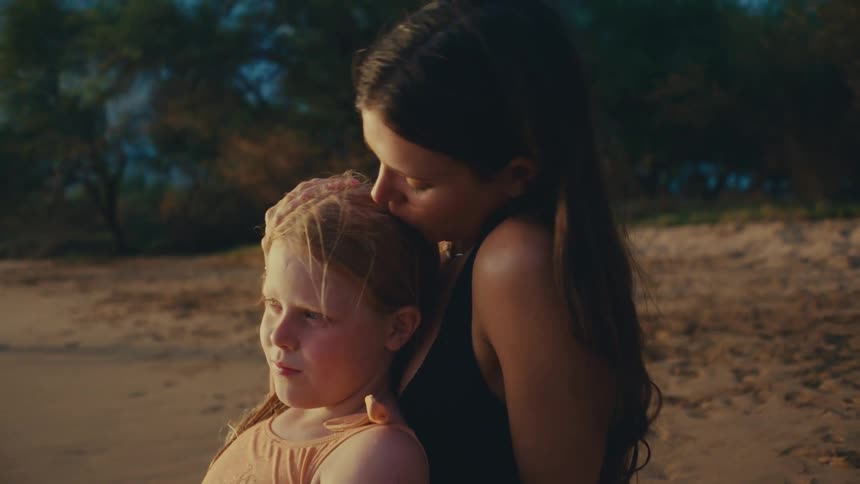 A mother sits on a beach with her daughter on her lap kissing her head under the soft sunlight of either a sunset or a morning