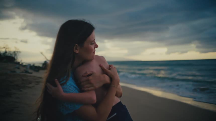 Mother with long dark hair wearing a light blue patterned topembraces and gazes affectionately at sonon a beach at dusk with the ocean and a cloudy sky in the background