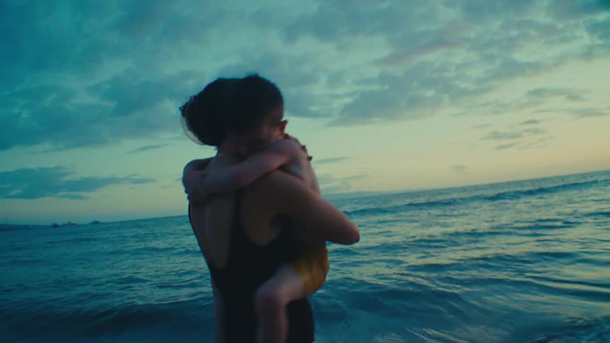 A mother in a dark swimsuit embraces her son in orange shorts on a beach at sunset with the ocean and a cloudy sky in the background