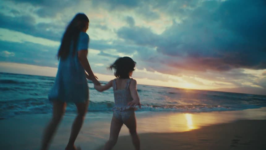 A woman and a young child run in silhouette on a beach at sunset with the ocean waves visible in the foreground