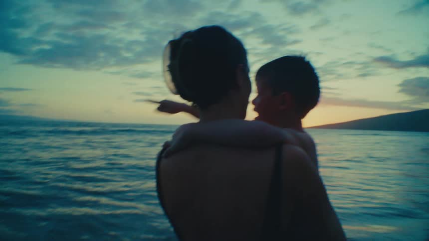 Mother holding her son in her arms while standing on the beach shoreline pointing towards the ocean during sunset