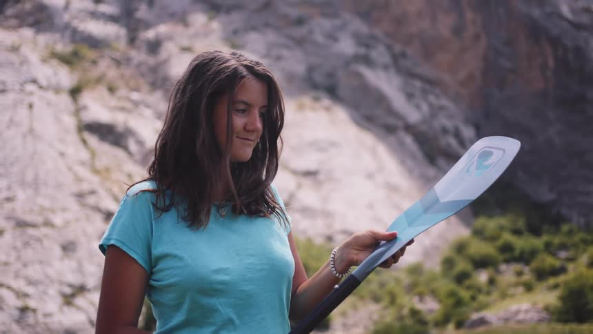A woman with long brown hair holds a blue paddle against a backdrop of rocky terrain