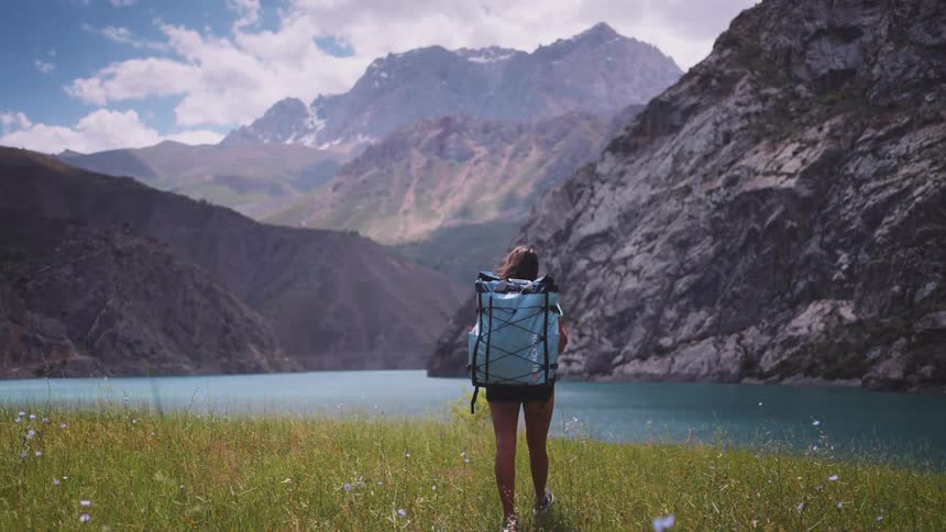 A woman carrying a backpack is hiking towards a lake by a rocky mountain range