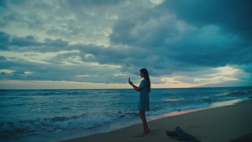 A woman stands at the edge of the water on a beach and takes pictures with her phone