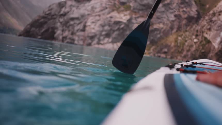 A paddle slices through clear blue lake water beside a vessel as it moves past towering rocky cliffs with ripples trailing from the blade and a hand visible gripping the side near tied gear