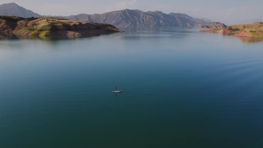 A person enjoys standup paddleboarding on a serene lake surrounded by mountains in the gentle light of morning